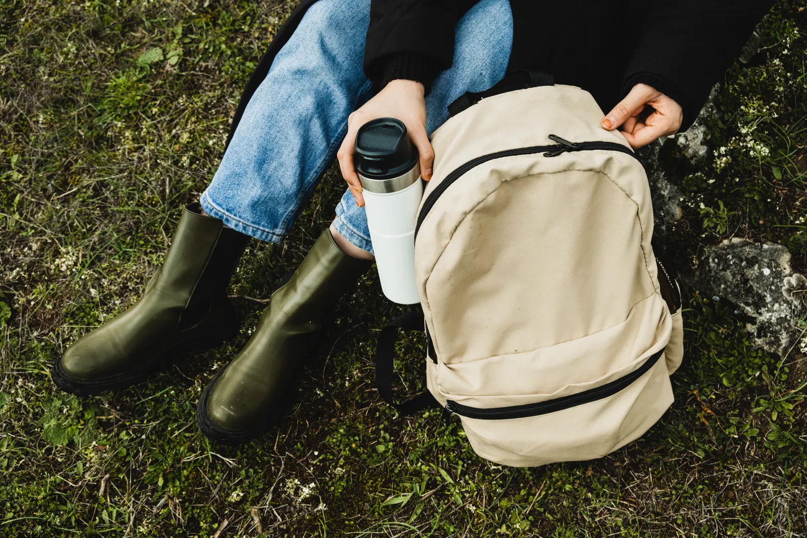 Person hält einen weißen Thermobecher neben einem Rucksack im Grünen