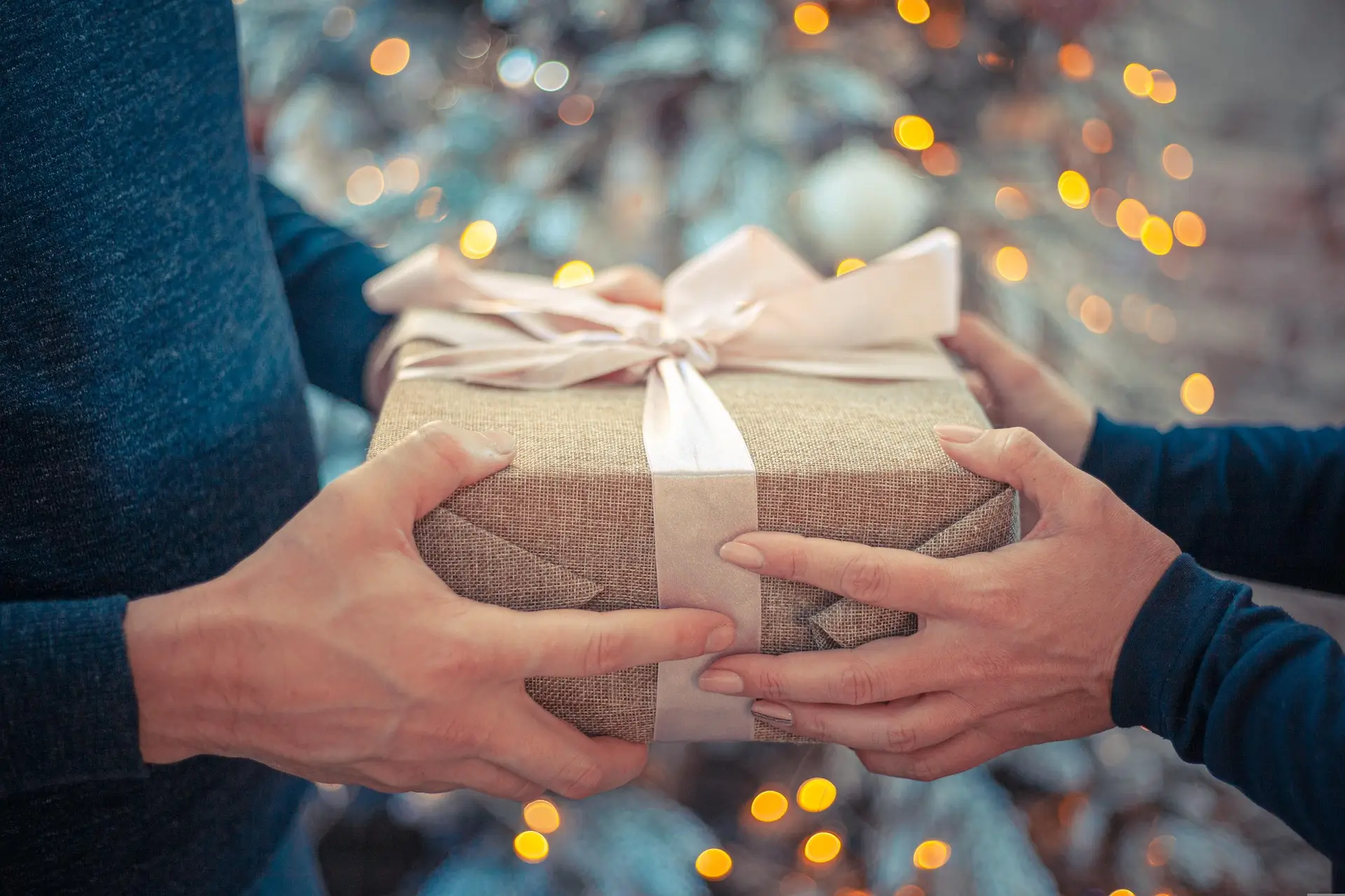 Zwei Personen reichen sich ein eingepacktes Geschenk mit weißer Schleife vor einem verschwommenen Weihnachtsbaum mit Lichtern