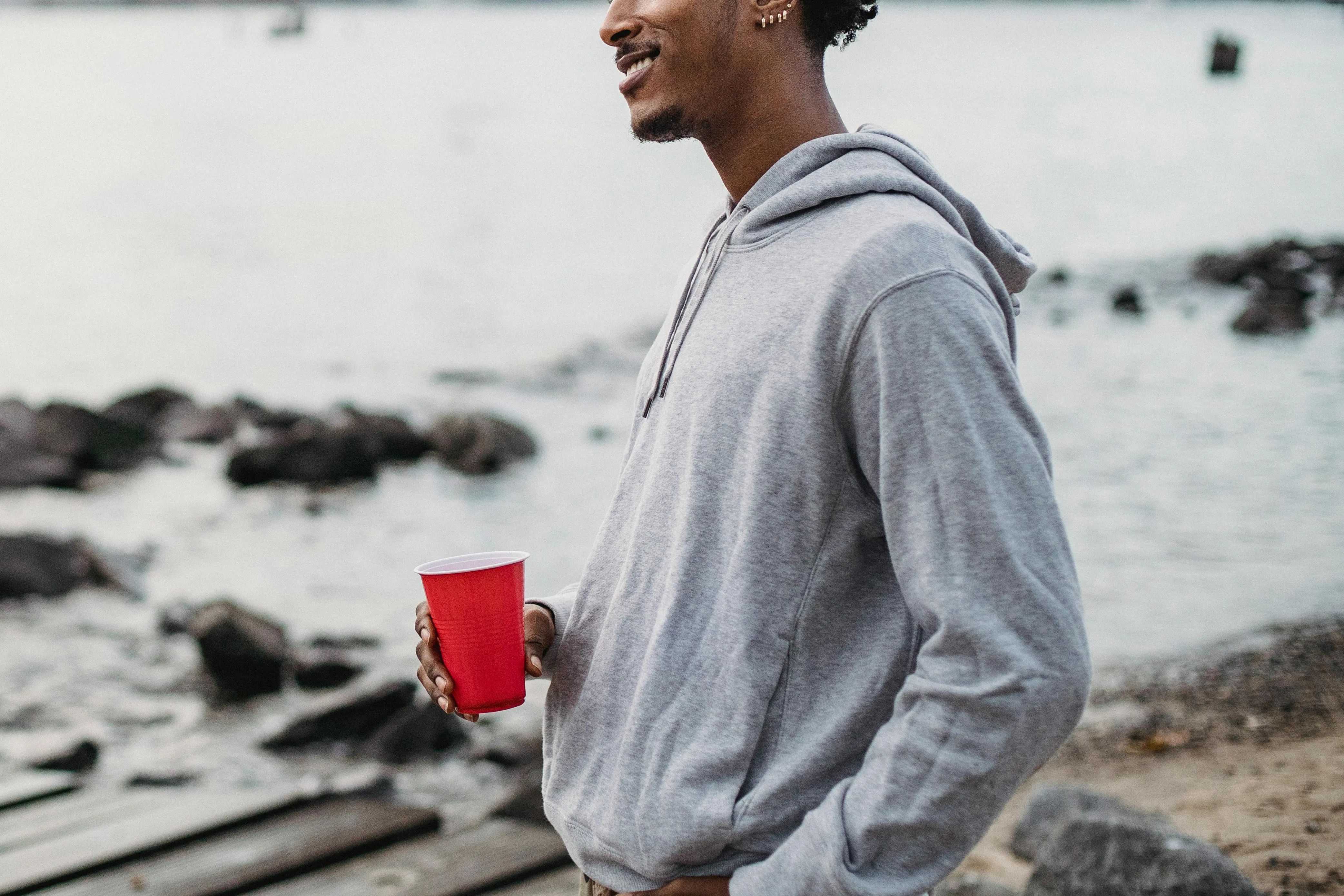 Person mit grauem Hoodie und rotem Becher am Ufer eines Sees