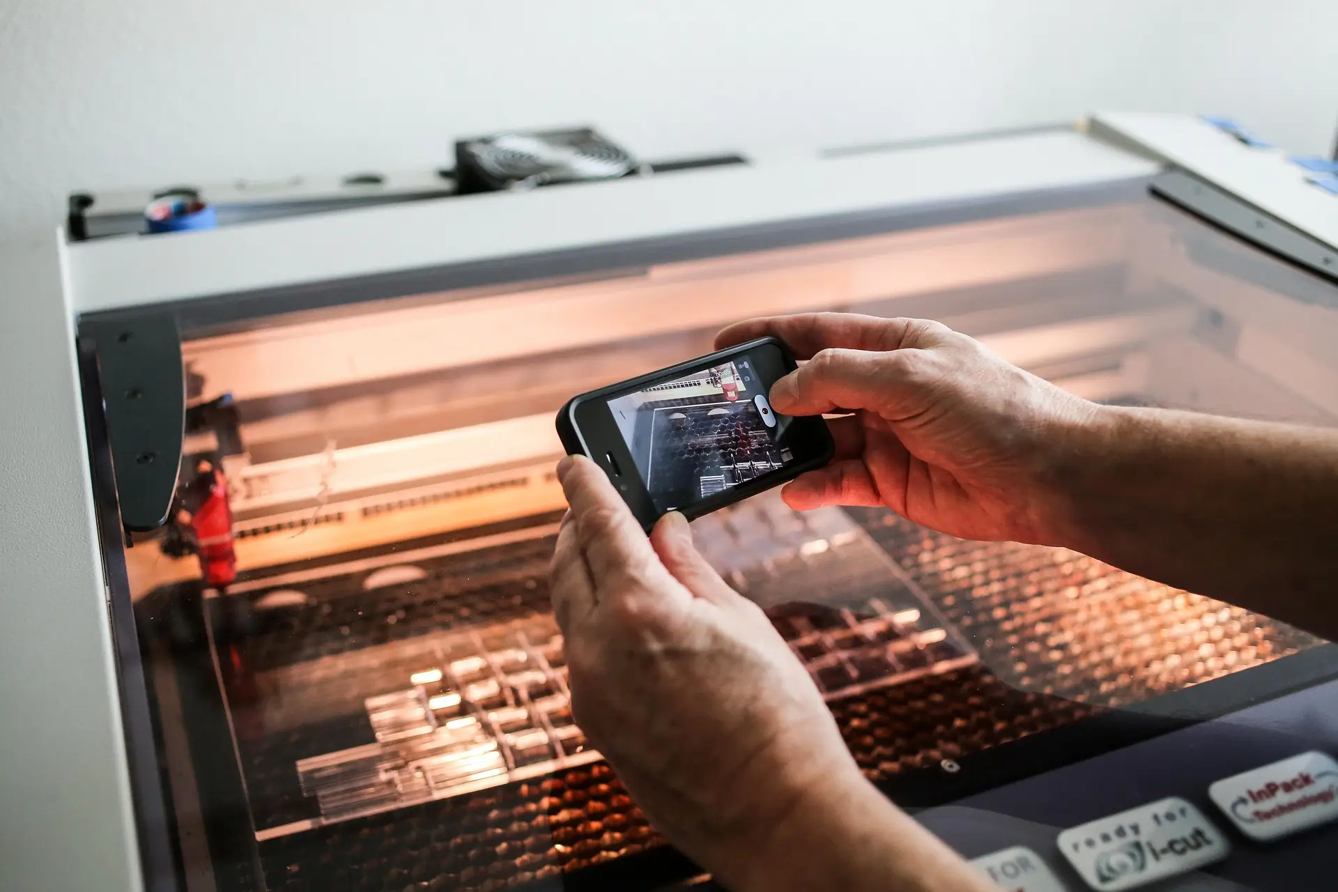 Hände halten ein Smartphone und fotografieren eine Laser Cutter Maschine mit beleuchtetem Arbeitsbereich in einer Werkstatt