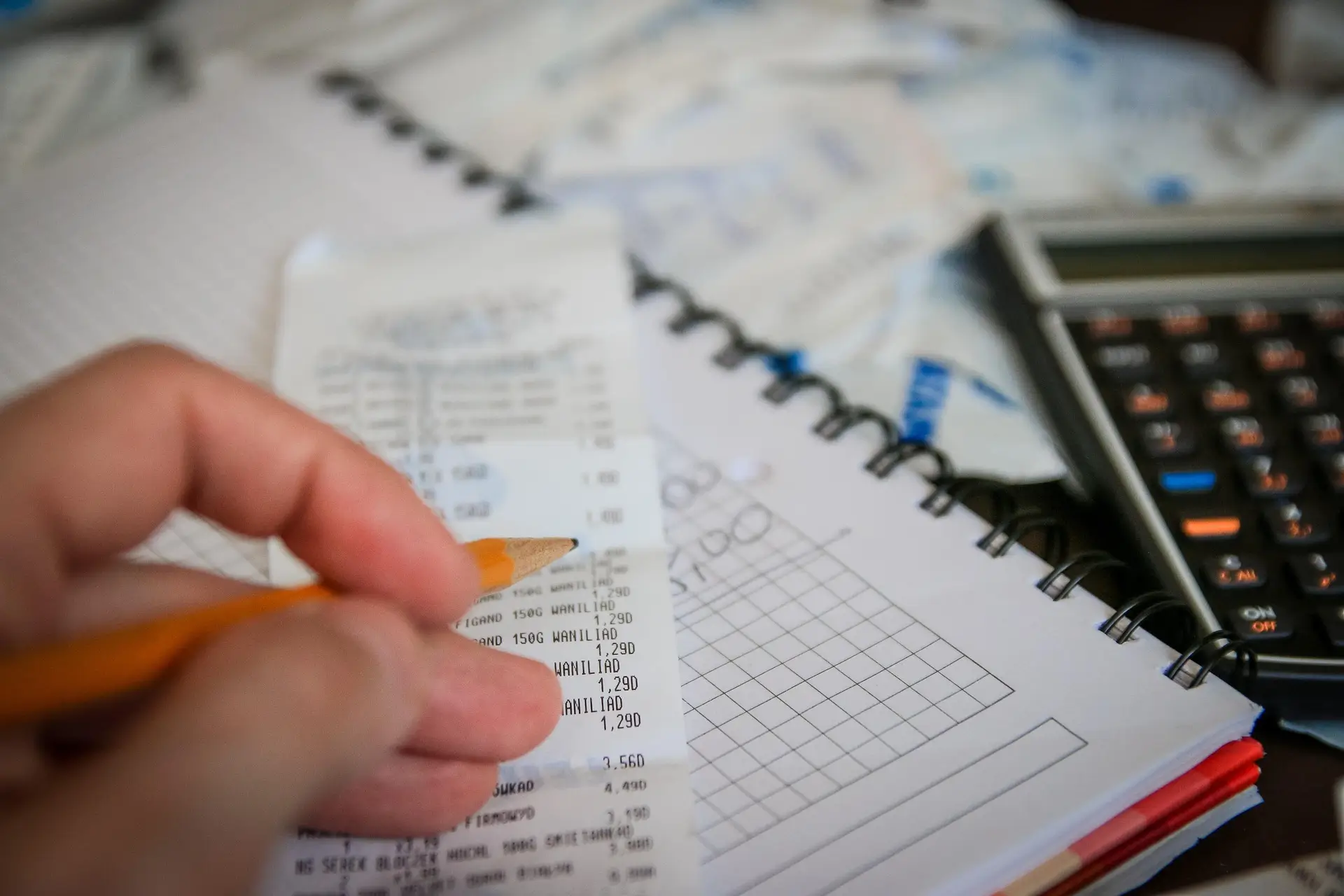 Hand hält einen Bleistift über Quittungen auf einem Notizbuch mit Tabelle, daneben liegt ein Taschenrechner im Hintergrund