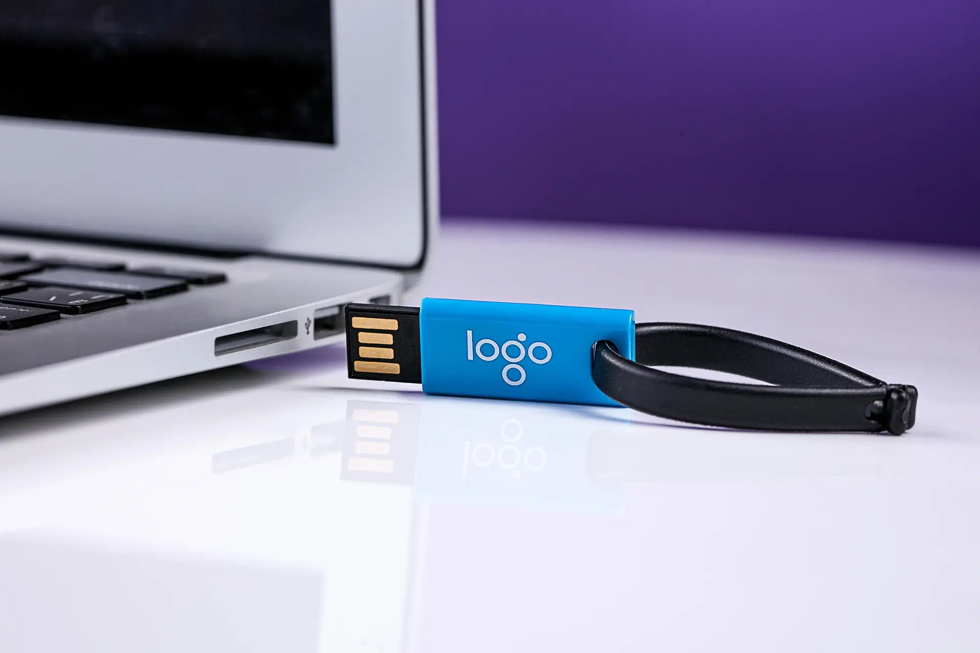 Blauer USB Stick mit Logo liegt neben einem Laptop und hat eine schwarze Schlaufe als Schlüsselanhänger