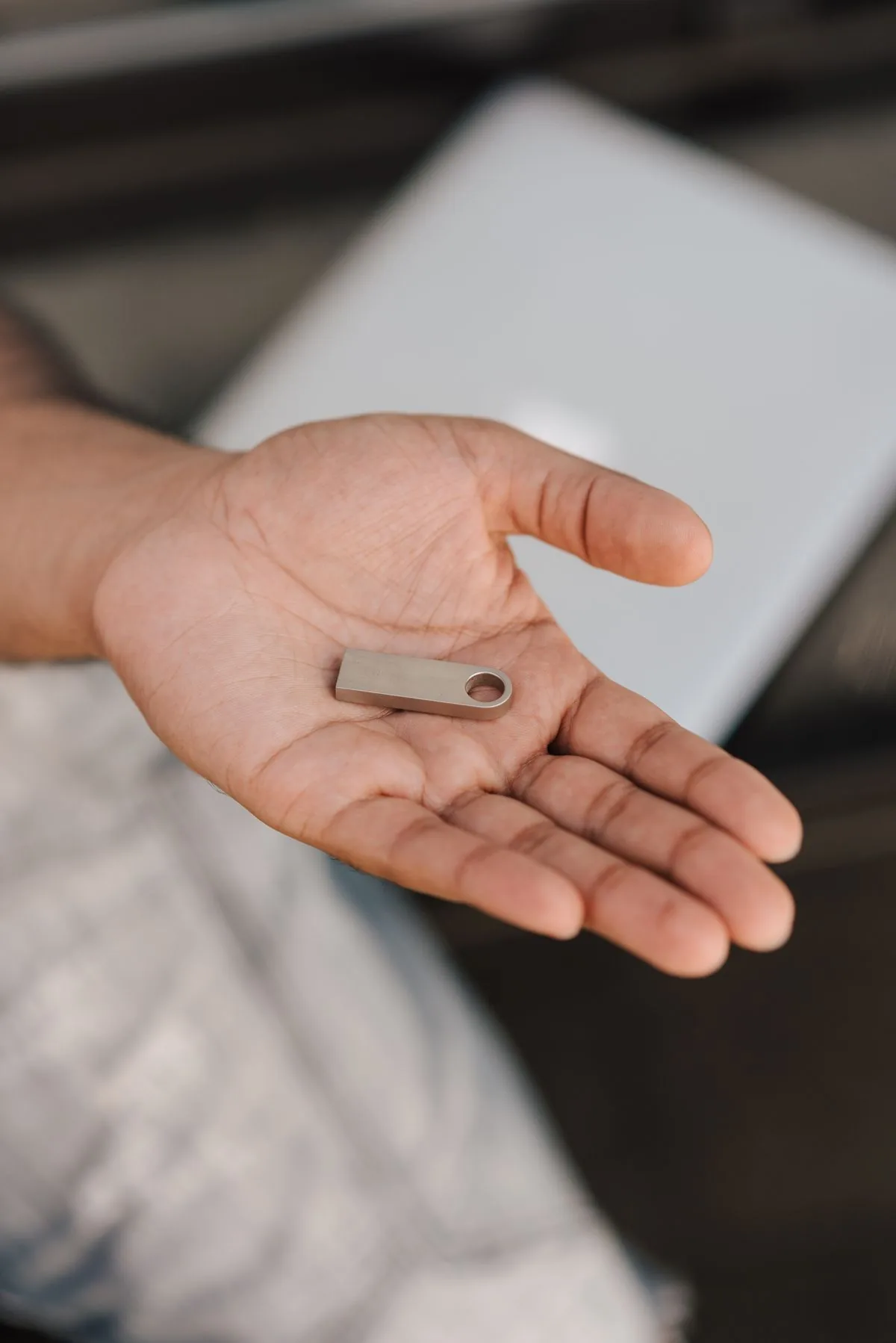 Hand hält einen silbernen USB-Stick, im Hintergrund ein Laptop