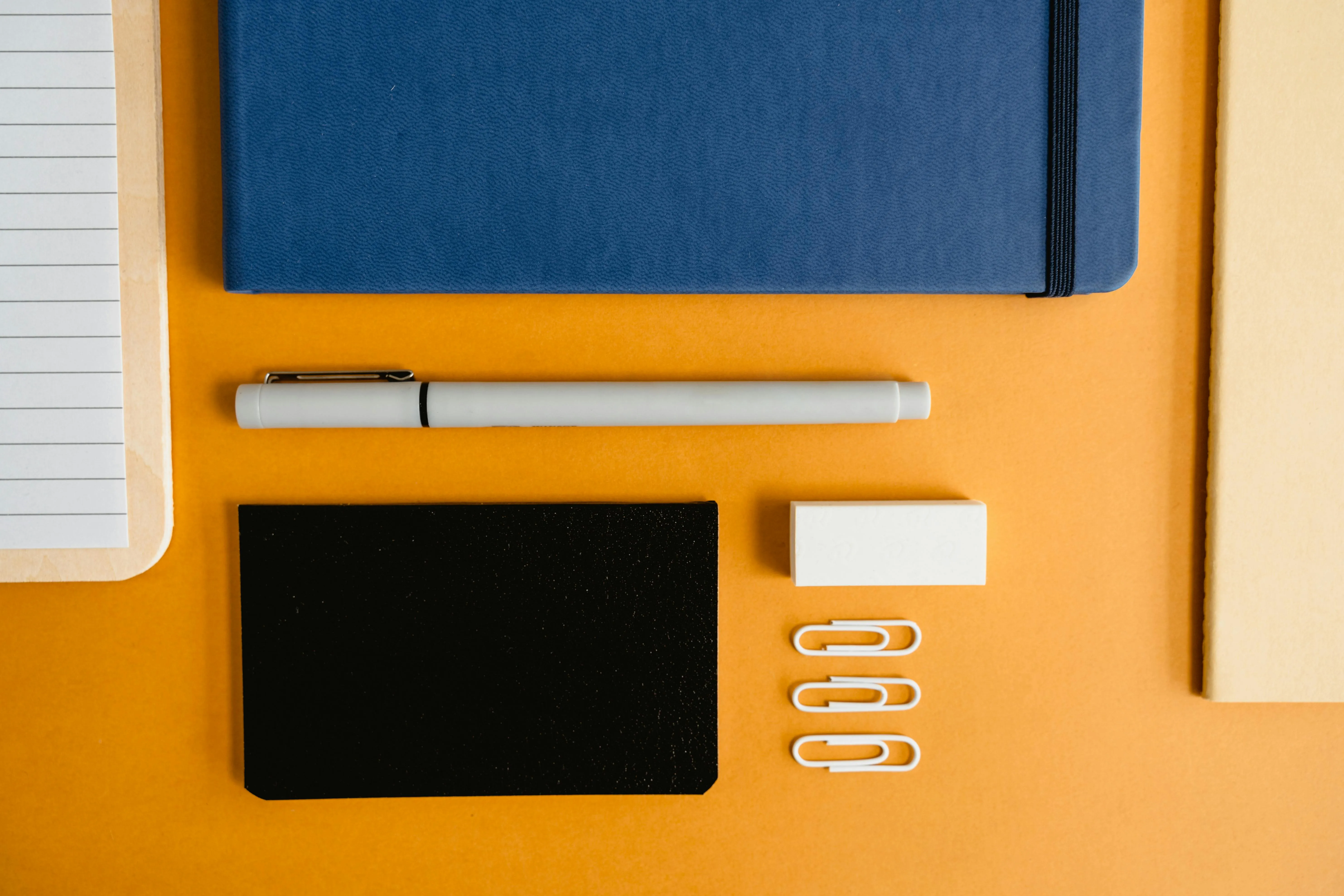 Notizbuch, Stift, Visitenkarten und Büroklammern angeordnet auf orangefarbenem Untergrund