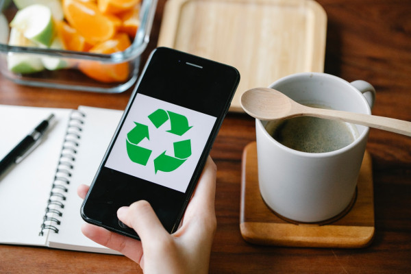 Hand hält ein Smartphone mit Recycling Symbol auf einem Schreibtisch neben Notizbuch, Kaffee und Obstbox