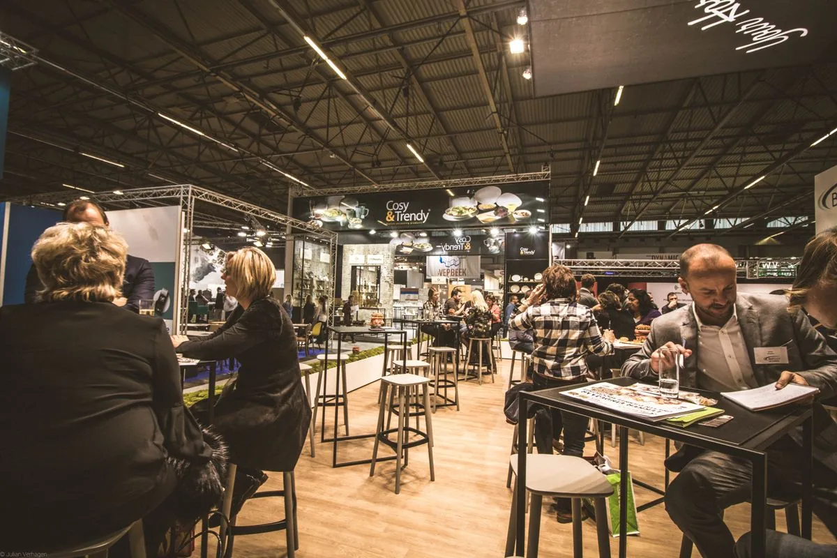 Besucher im Gespräch an Stehtischen zwischen beleuchteten Messeständen in einer Messehalle