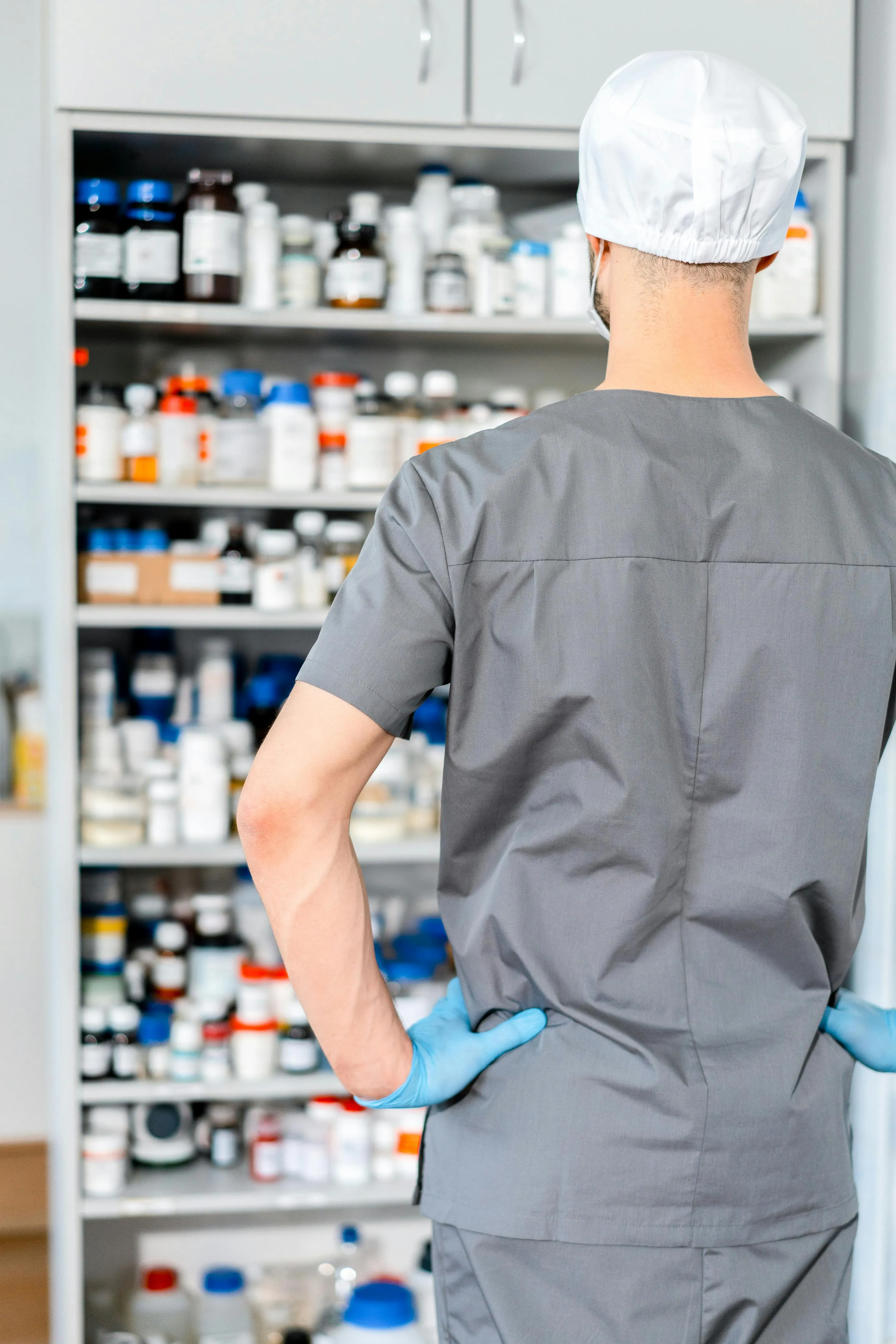 Apotheker in grauer Arbeitskleidung und OP-Haube steht mit dem Rücken zur Kamera vor einem gefüllten Medikamentenschrank.
