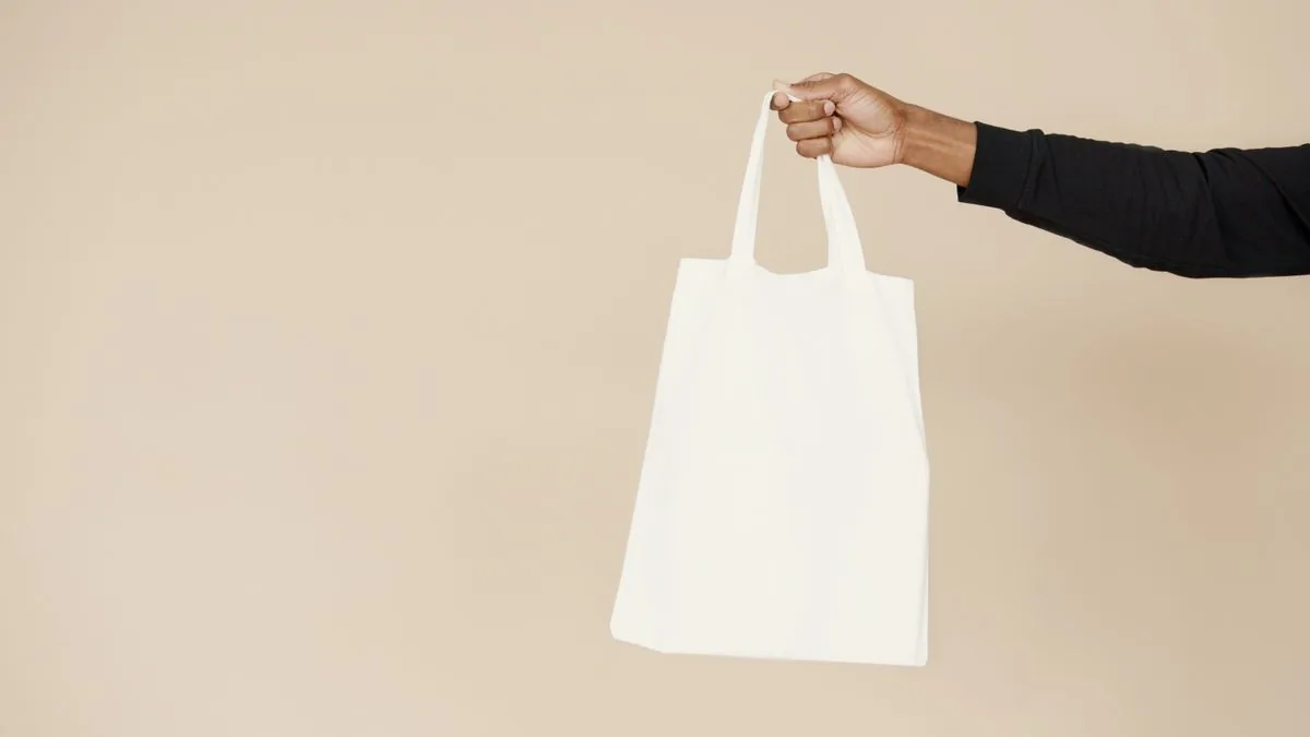 Hand hält eine unbedruckte weiße Baumwolltasche vor beigefarbenem Hintergrund